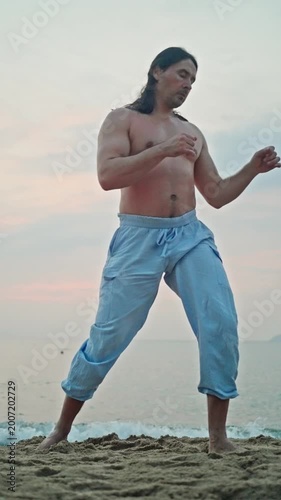 Sunset At Coast With Fluid Movements. Male Practicing Rhythmic Kick Sequences By Seaside During Sunset. Calm Coastal Scene With Barefoot Man Engaged In Expressive Kicking Routines At Sunset