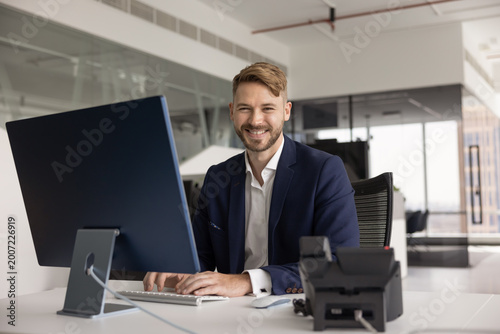 Foto Portrait of smiling confident businessman in suit sits at desk in modern office, looking at camera while typing on computer keyboard