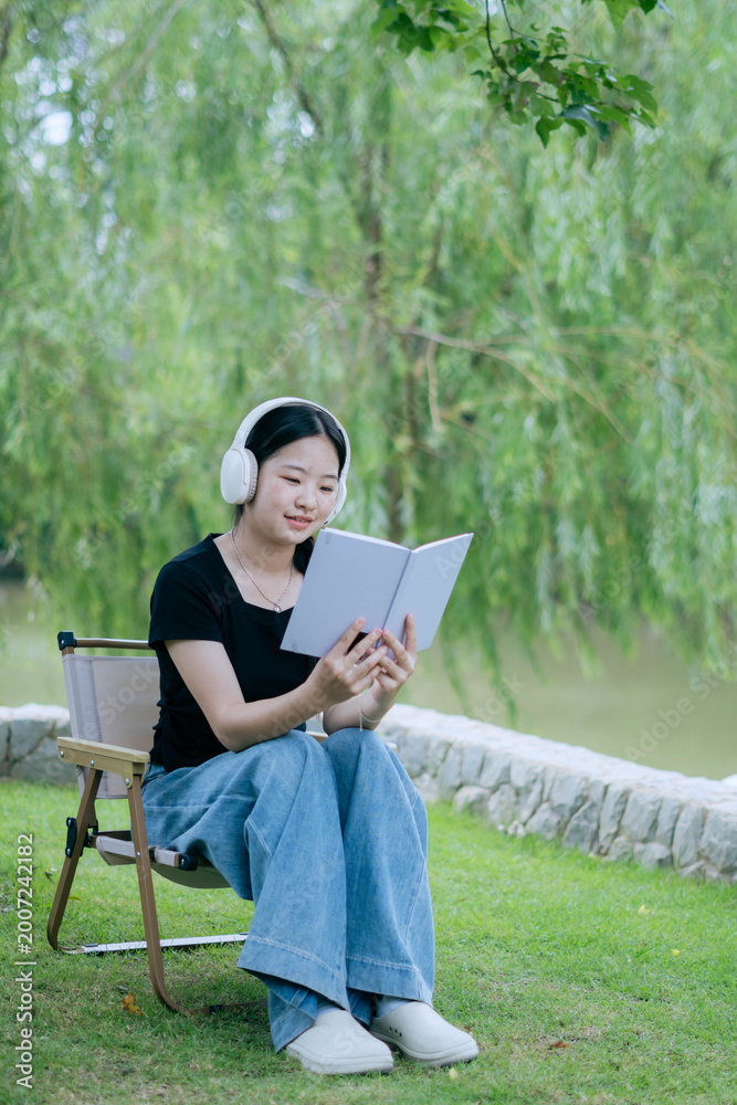 Fototapeta premium Young Woman Reading Book with Headphones in Nature Park