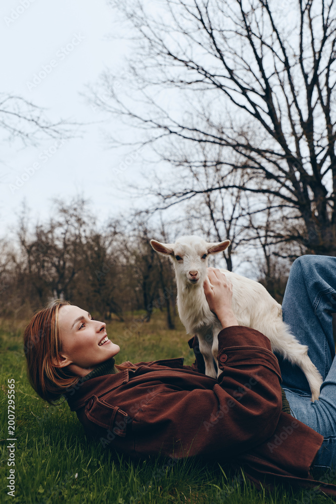 Fototapeta premium Relaxed woman lies on grass in a park, stroking a playful goat kid, enjoying outdoor life and gentle connection with nature, candid moment of simple countryside joy