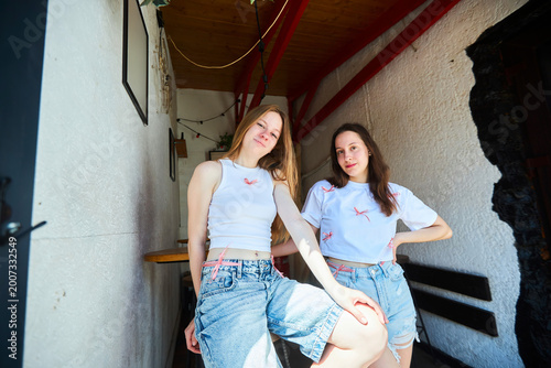 Two beautiful young girls in a white top and denim shorts are sitting at a table in a cafe. Fashion, beauty and youth. The time of the year is summer