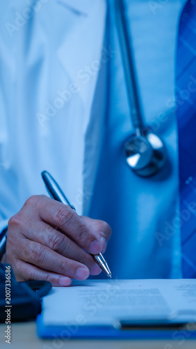 Male doctor wearing white lab coat stethoscope writes medical document upon clipboard inside blue clinical office setting performing daily healthcare duties hospital staff clinic desk patient record.