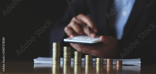 Businessman calculating finances with stacked coins and calculator, representing budgeting, financial analysis, and business accounting