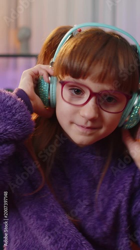 Happy overjoyed child girl kid in wireless headphones relaxing sitting on couch at night home choosing listening favorite energetic disco dancing music. People weekend leisure activities. Vertical