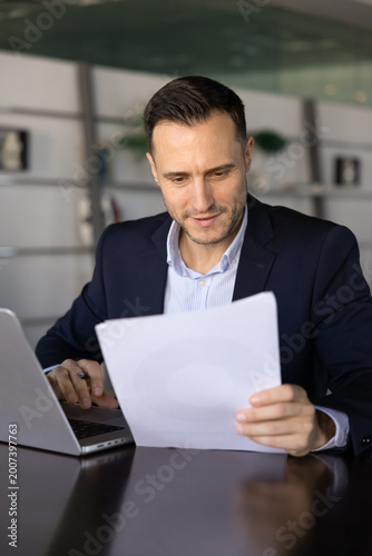 Focused adult businessman sits at desk in office, carefully studying printed documents while working on laptop, represents analysis, decision-making, business materials and contract details review