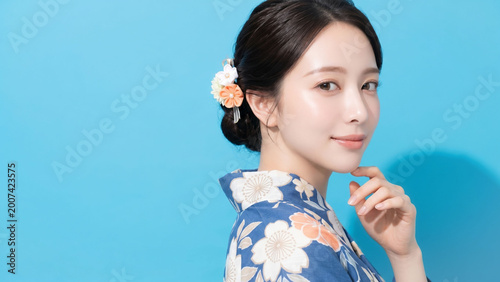 鮮やかなブルー背景の前で、花柄の青い浴衣と可憐な髪飾りをまとい、やさしく微笑みながら振り返る若い女性の和風ビューティーポートレート。夏祭り、和装、観光、伝統文化、美容、季節キャンペーン、旅行プロモーションなど幅広い広告用途に使いやすい、爽やかで上品な印象の人物イメージ素材