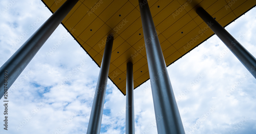 Fototapeta premium Abstract Worm's Eye View of Modern Architectural Pillars Supporting a Vibrant Yellow Canopy Against a Cloudy Blue Sky in Budapest
