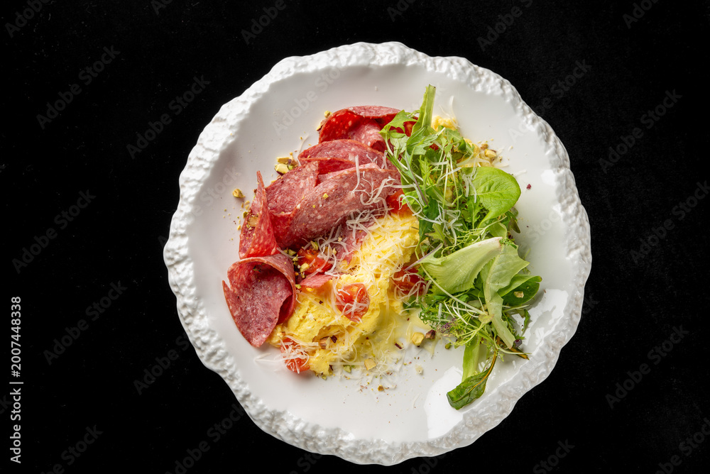 Obraz premium Gourmet omelet served with salami, cherry tomatoes, and a fresh green salad on a textured white plate. Dark background, professional food photography for restaurant menu and culinary concepts