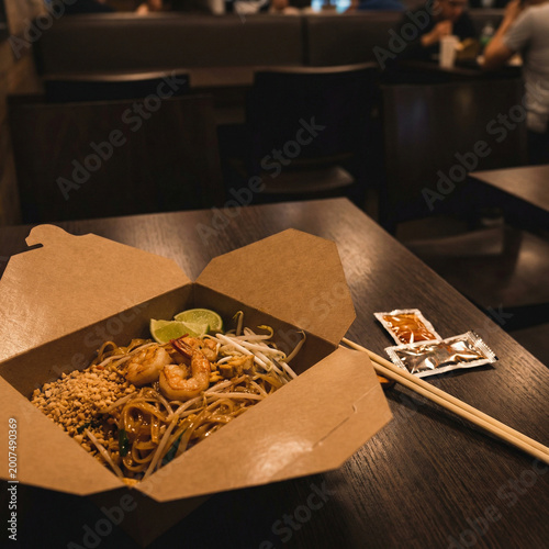 Shrimp noodle takeout box with lime wedge, bean sprouts, and crushed peanuts on a wooden table in a restaurant interior, emphasizing casual dining and ready-to-eat Asian food service