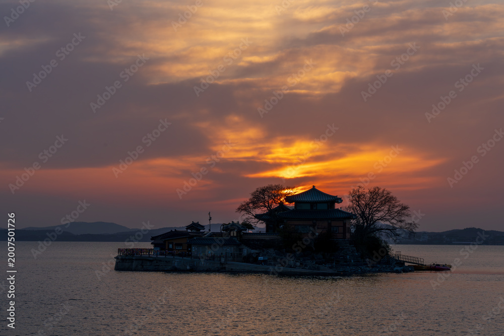 Obraz premium Serene sunset view of Ganworam Hermitage, a unique Buddhist temple on a tiny island in Seosan, South Korea, with the glowing sun and vibrant orange sky reflected on the sea.