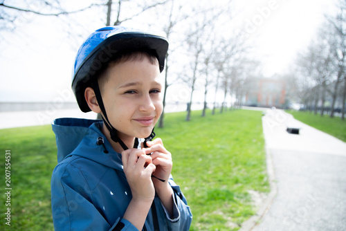Close-up of a boy of European appearance in a blue jacket with a blue protective helmet on his head smiles at the camera against the background of the lawn and trees. Concept of safety rules, personal