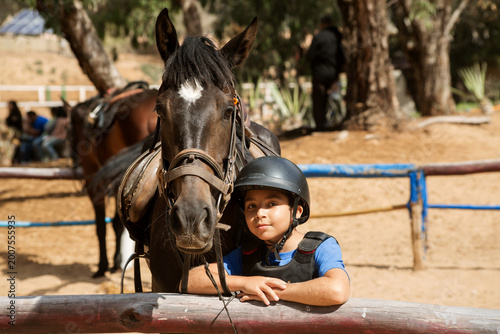 Portrait of a young boy in a riding helmet and protective vest standing next to a saddled horse at an outdoor stable. T
