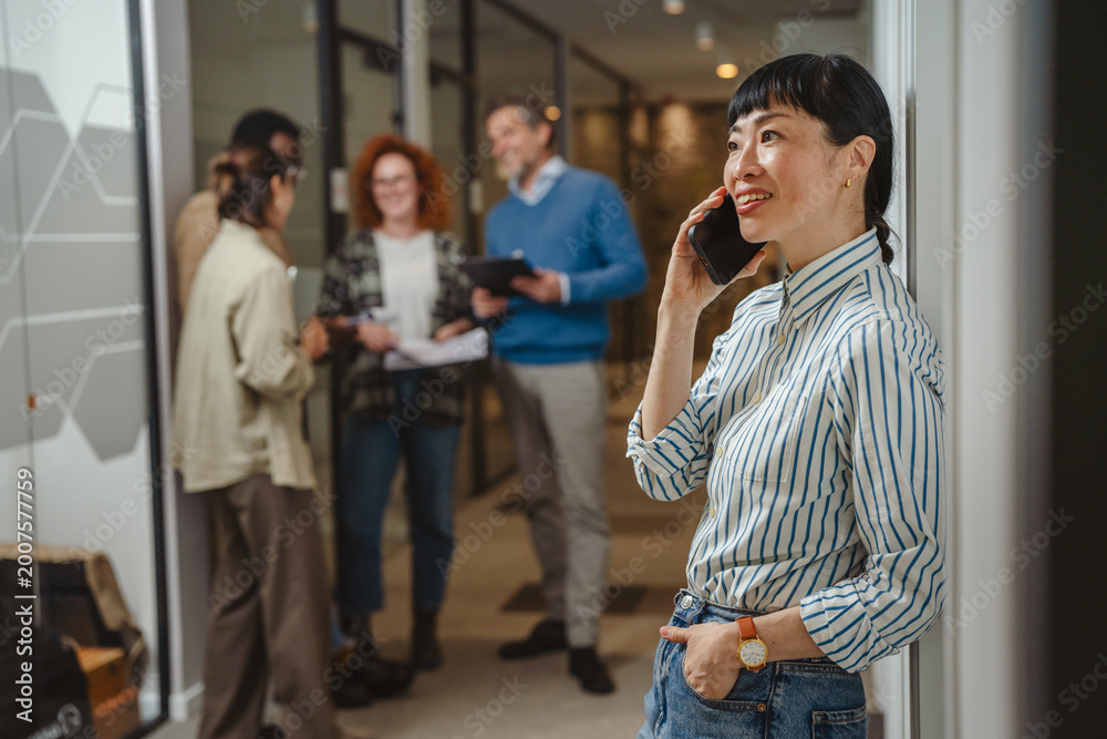 Fototapeta premium Woman talking on smart phone in office hallway with colleagues