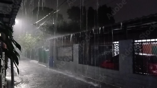 Heavy rain falling on a residential street at night with visible water splashes and dim lighting, creating a moody urban atmosphere