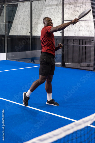 Male athlete swinging padel racket on blue court with glass walls, mesh netting and doodle panel