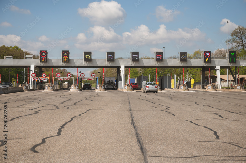 Obraz premium Multi lane toll road entrance with electronic signs showing speed limits and fees, vehicles approaching payment booths on busy highway, road infrastructure and commuter transport concept