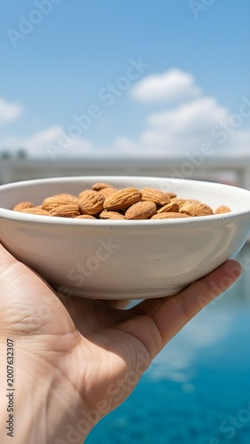 Hand Holding a Bowl of Raw Almonds Outdoors with Blue Sky and Pool Background