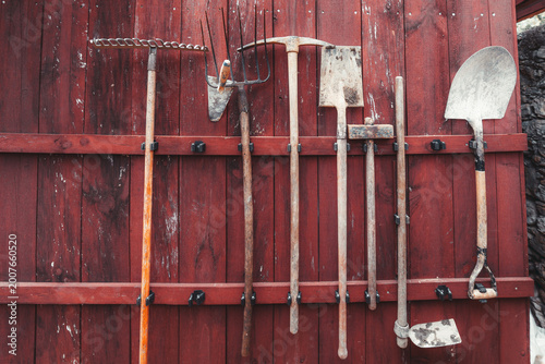 Vintage garden tools hanging on a weathered red wooden wall, with rake, shovel, hoe, pickaxe and fork arranged in rustic farm storage