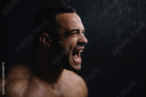 Wet muscular man screaming emotional expression dramatic lighting water droplets dark background intense portrait strength determination cinematic style vertical composition