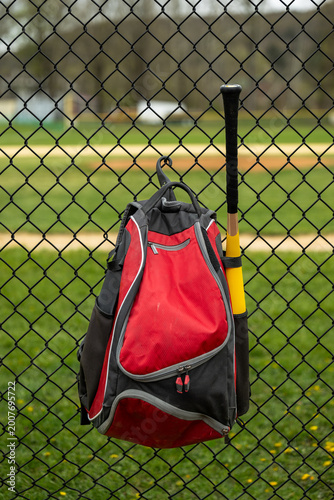 фотографія A red baseball equipment bag with a bat hangs from the chain link fence at a youth baseball field, the green grass of the outfield visible