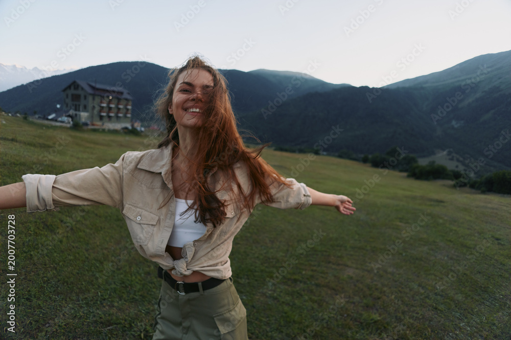 Naklejka premium Cheerful woman outdoors, embracing a sunlit moment in a green field, arms open, carefree smile, natural breeze, mountain backdrop, vibrant natural colors with warmth.