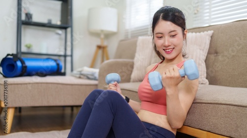 Asian young women exercise using dumbbells lifting weights home workout fitness training sitting couch living room healthy lifestyle wellness indoor physical