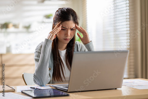 Frustrated Asian woman experiences severe headache and stress while working on laptop computer at home office desk feeling overwhelmed and exhausted from excessive screen time and heavy workload