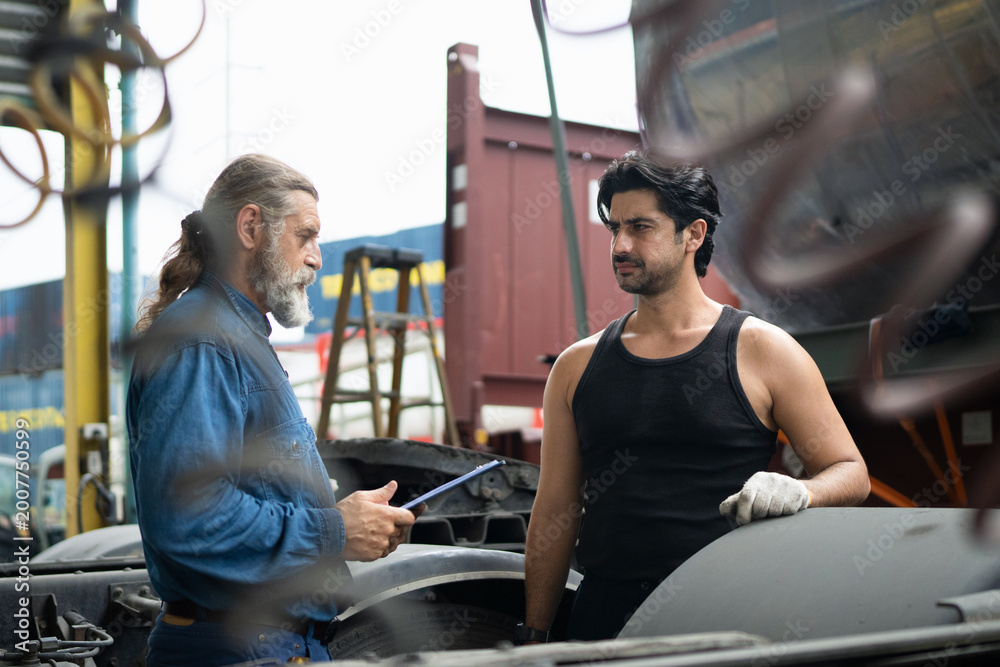Fototapeta premium Two mechanics collaborating on truck maintenance, with a supervisor inspecting the work at an industrial vehicle depot to ensure quality control and safety
