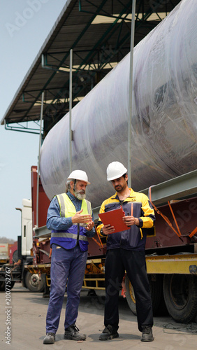 Two construction workers walking and discussing safety near trucks on site wearing helmets and uniforms. two male using a tablet beside a truck trailer and stacked cargo containers