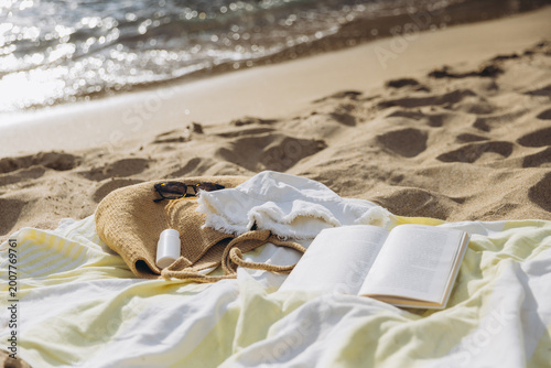 Billede på lærred Beach essentials on a sandy shore by the ocean, with an open book, straw bag, an