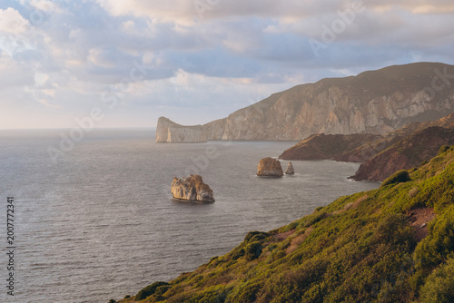 Sardinia coastline showing the sea stack Pan di Zucchero, dramatic cliffs, and green hills bathed in soft sunset light