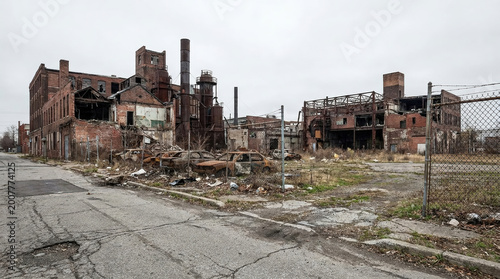 Old factory buildings sit abandoned on a dusty street with weeds and debris scattered around under a gray sky on a chilly day in an industrial area
