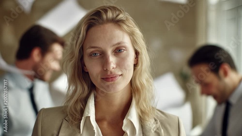 A blonde woman in an office, surrounded by colleagues throwing papers at each other, remains completely calm. Stress resistance, project completion, composure.