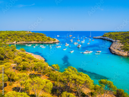 Aerial view of luxury yachts and boats anchored in a turquoise bay with crystal clear water on the coast of Mallorca, Spain. Scenic Mediterranean landscape during a bright sunny summer day.