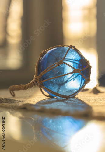 Photography Vertical composition of blue glass fishing float with rope net and reflection in warm indoor light setting