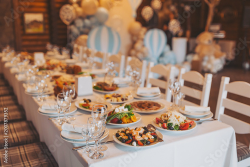 A long table is arranged with numerous plates of food and drinks. The setting features balloons and decorations, creating a festive atmosphere. Guests will soon enjoy the meal together