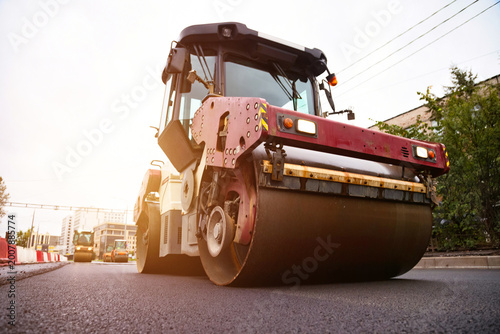Heavy machinery is used to lay asphalt concrete on a busy city street. Road construction is underway, improving infrastructure and traffic flow. The sun sets behind the vehicles.