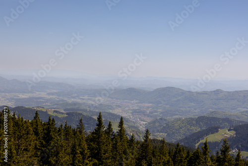 High-angle panoramic view of a vast mountain valley with rolling hills, distant hazy peaks, and dark evergreen treetops in the foreground under a clear sky.