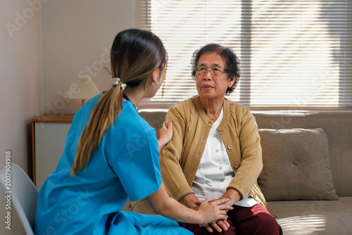 Professional caregiver, wearing blue scrubs, comforting an elderly Asian woman sitting on a sofa in a bright living room, providing support
