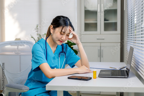 Tableau sur toile Young Asian nurse working on laptop at desk, experiencing stress, fatigue, and h