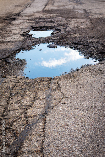 Obraz na płótnie A large, water-filled pothole on a cracked asphalt road reflects a bright blue sky and white clouds, emphasizing road deterioration