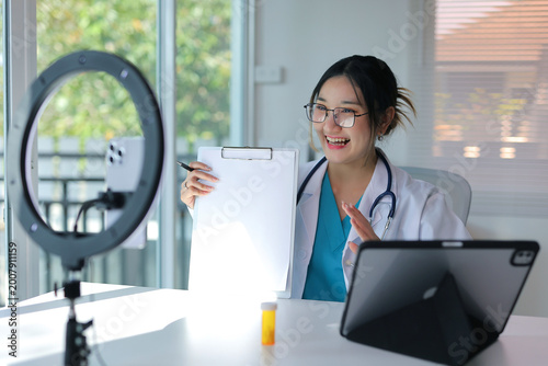 Female physician in white coat consulting virtually, presenting information on a clipboard during a telehealth video conference
