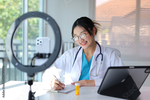 Female doctor in lab coat and stethoscope consulting patients remotely while making notes, using phone and tablet for digital healthcare