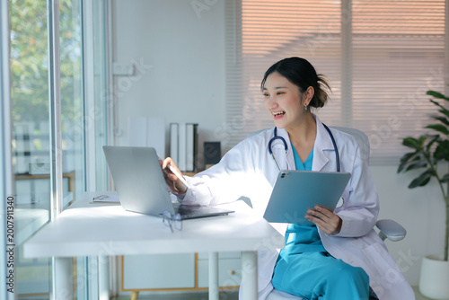 Smiling female doctor wearing a lab coat and stethoscope, working on a laptop and holding a digital tablet in a modern office