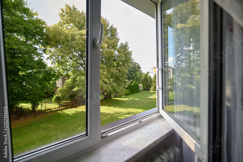 Fotografie Open white PVC window overlooking a green residential garden with mature trees and a mown lawn, Warsaw, Masovian, Poland, Europe