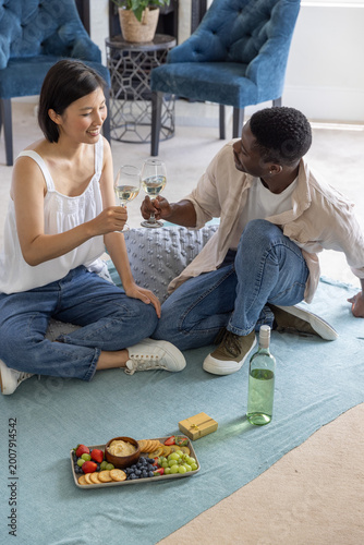 Diverse couple clinking wine glasses, sharing snack platter, sitting on light-blue rug in home