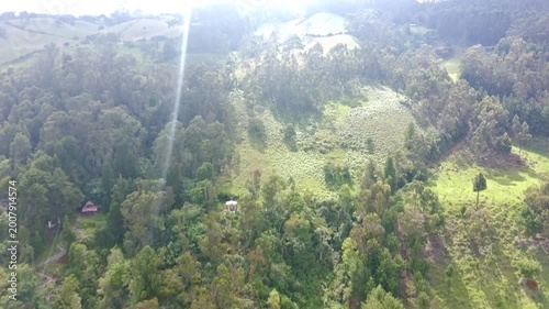 High-angle aerial shot of a dense green forest and sloping hillsides under a bright sky with light mist on the horizon.