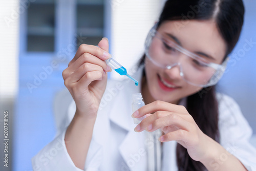 Fotografie Young Asian scientist in lab coat and safety glasses dropping blue liquid from a