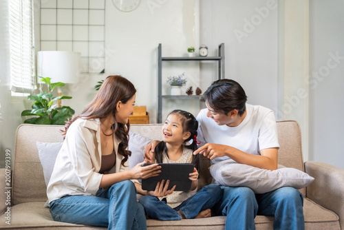 Asian parents and daughter sitting on a couch, watching content and interacting with a digital tablet at home