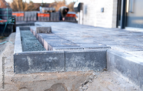Construction site featuring newly laid gray concrete pavers forming a patio area with a worker in the background arranging additional pavers and tools visible